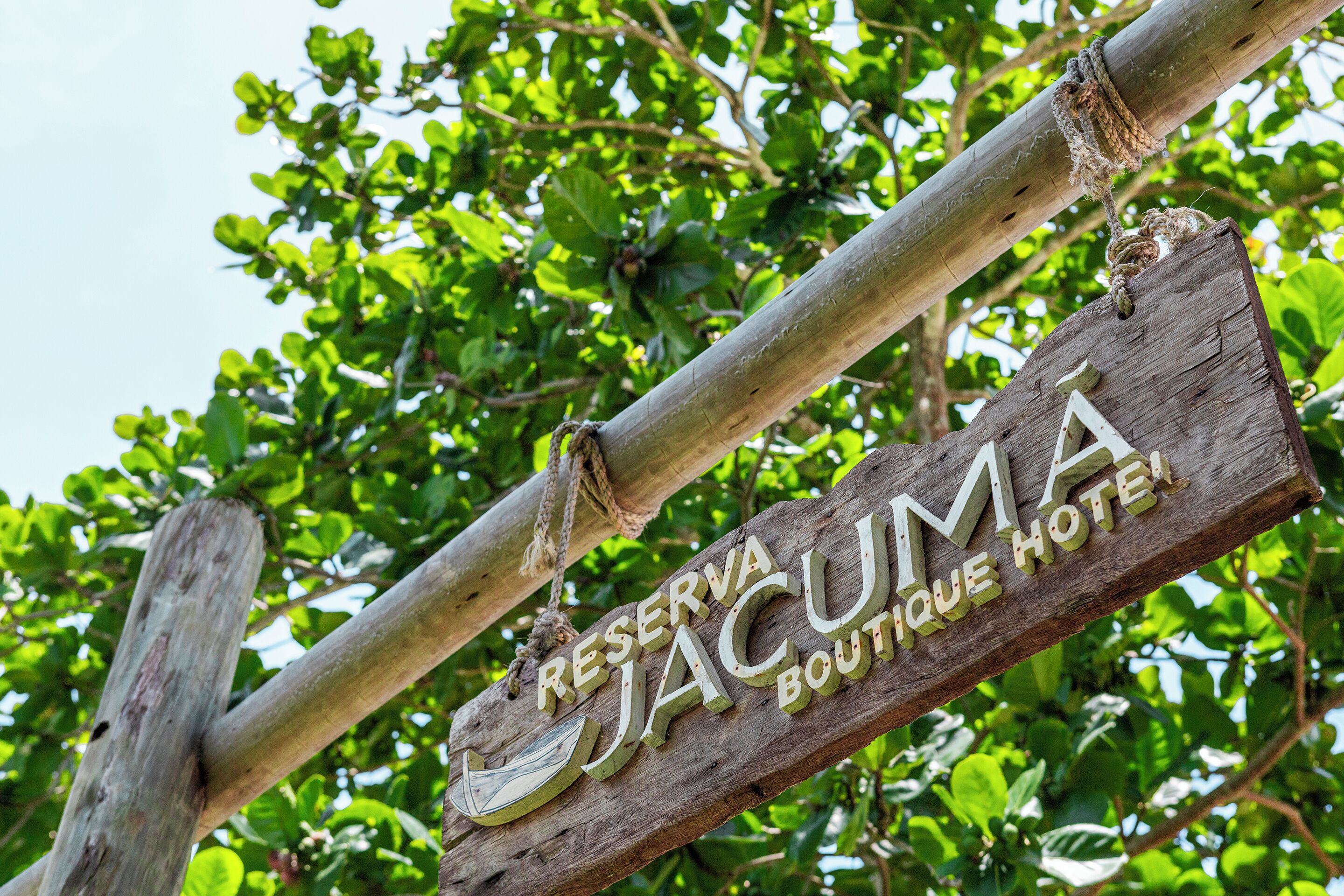 A wooden sign with the text "RESERVA JACUMA BOUTIQUE HOTEL" hanging from a tree branch.