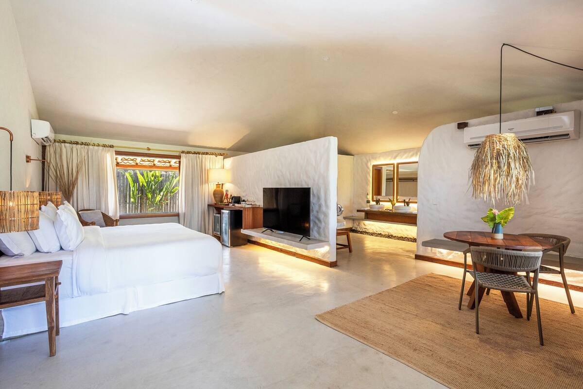 A spacious hotel room with a large bed, a TV, a dining area with a table and chairs, and a small kitchen area.