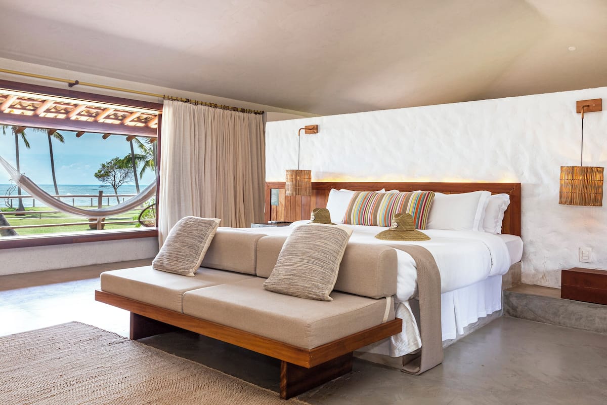 A bedroom with a large bed, a sofa, a view of the beach, and a hammock outside.