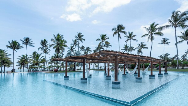A large, clear swimming pool with a wooden pergola and palm trees in the background. - Reserva Jacumã Boutique Hotel  (Porto Seguro)