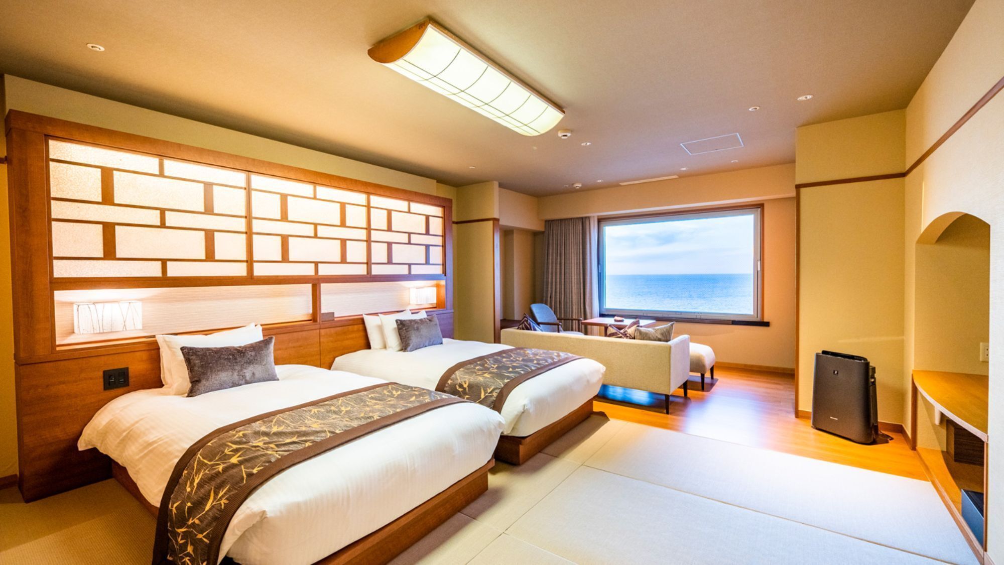 A hotel room with two beds, a desk, a chair, a TV, and a large window with a view of the sea.