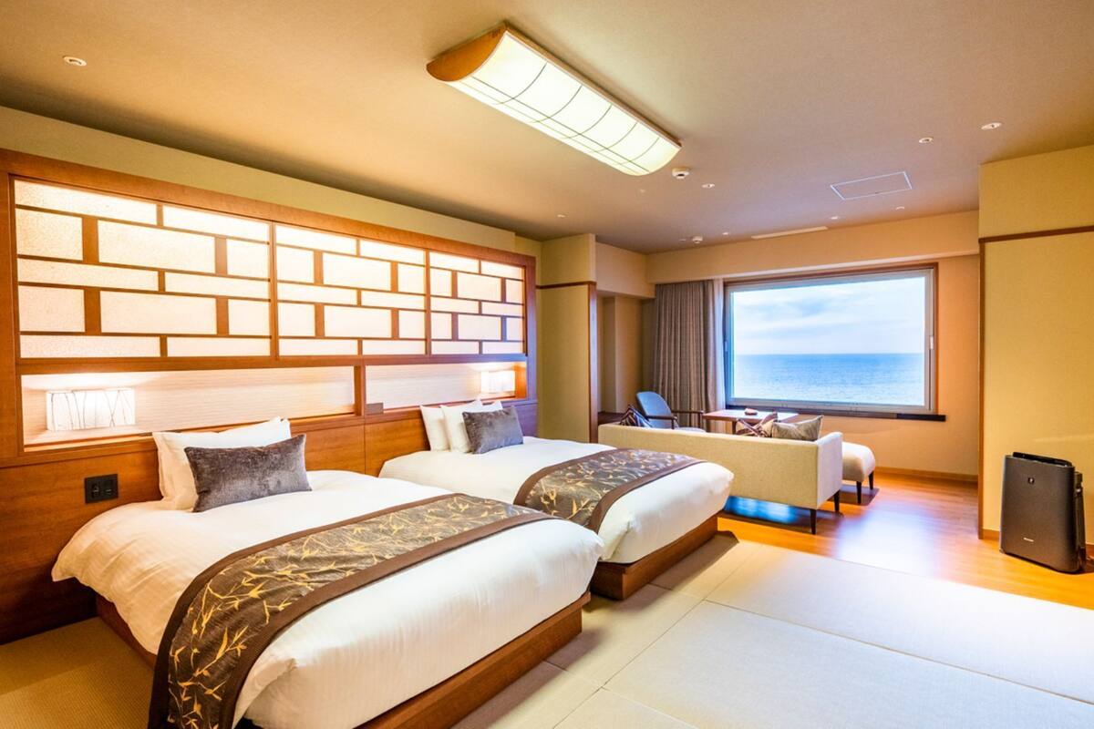 Une chambre d’hôtel avec deux lits, un bureau, une chaise, une télévision et une grande fenêtre donnant sur la mer.