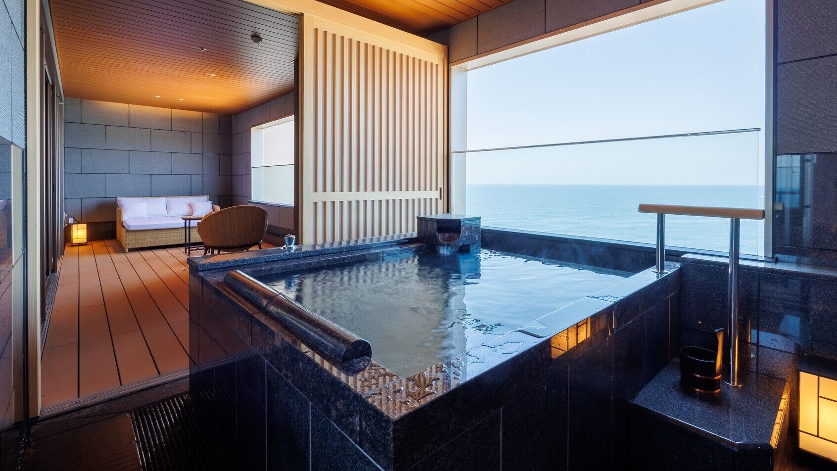 A modern indoor pool with a view of the sea, a wooden deck, and a seating area with a sofa and chairs.