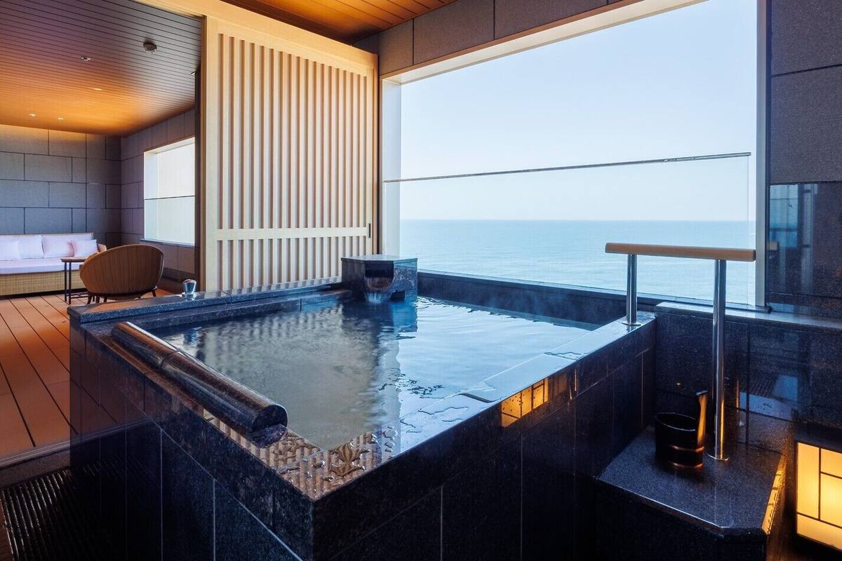 Une piscine intérieure moderne avec vue sur la mer, une terrasse en bois et un espace détente avec un canapé et des fauteuils.