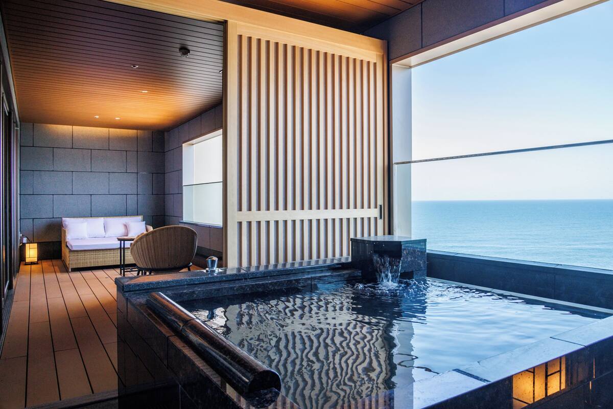 Un espace piscine intérieur moderne avec vue sur la mer, un coin salon avec un canapé et des fauteuils, et un plancher en bois.