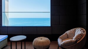 Una habitació amb un gran finestral amb vista al mar, una cadira de vimet, una taula rodona petita i un sofà.
