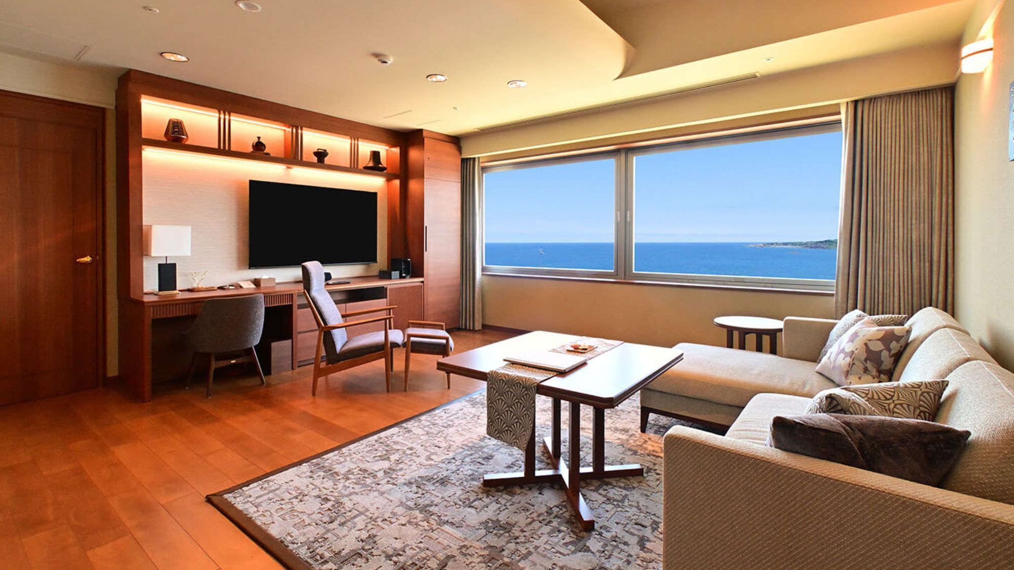 A modern hotel room with a large window offering a sea view, a cozy seating area, a flat-screen TV, and a desk with a chair.