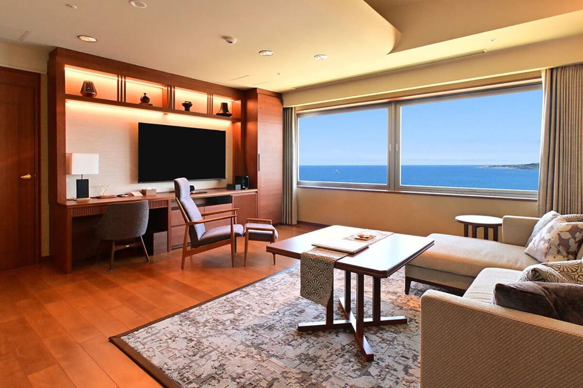 Une chambre d’hôtel moderne dotée d’une grande fenêtre donnant sur la mer, d’un coin salon confortable, d’une télévision à écran plat et d’un bureau avec une chaise.