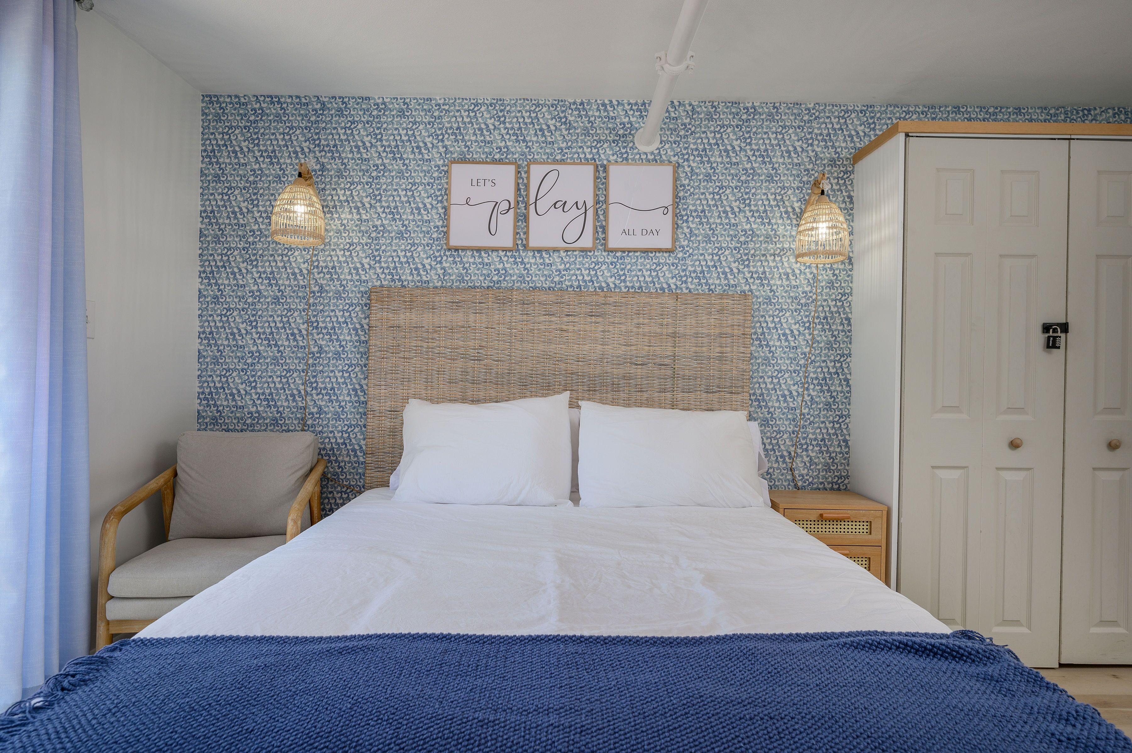 Un lit bien fait, avec une couverture bleue, une tête de lit en osier et un mur recouvert d’un papier peint à motifs bleus. On y trouve deux lampes fixées au mur et une petite table de chevet en bois avec une lampe.
