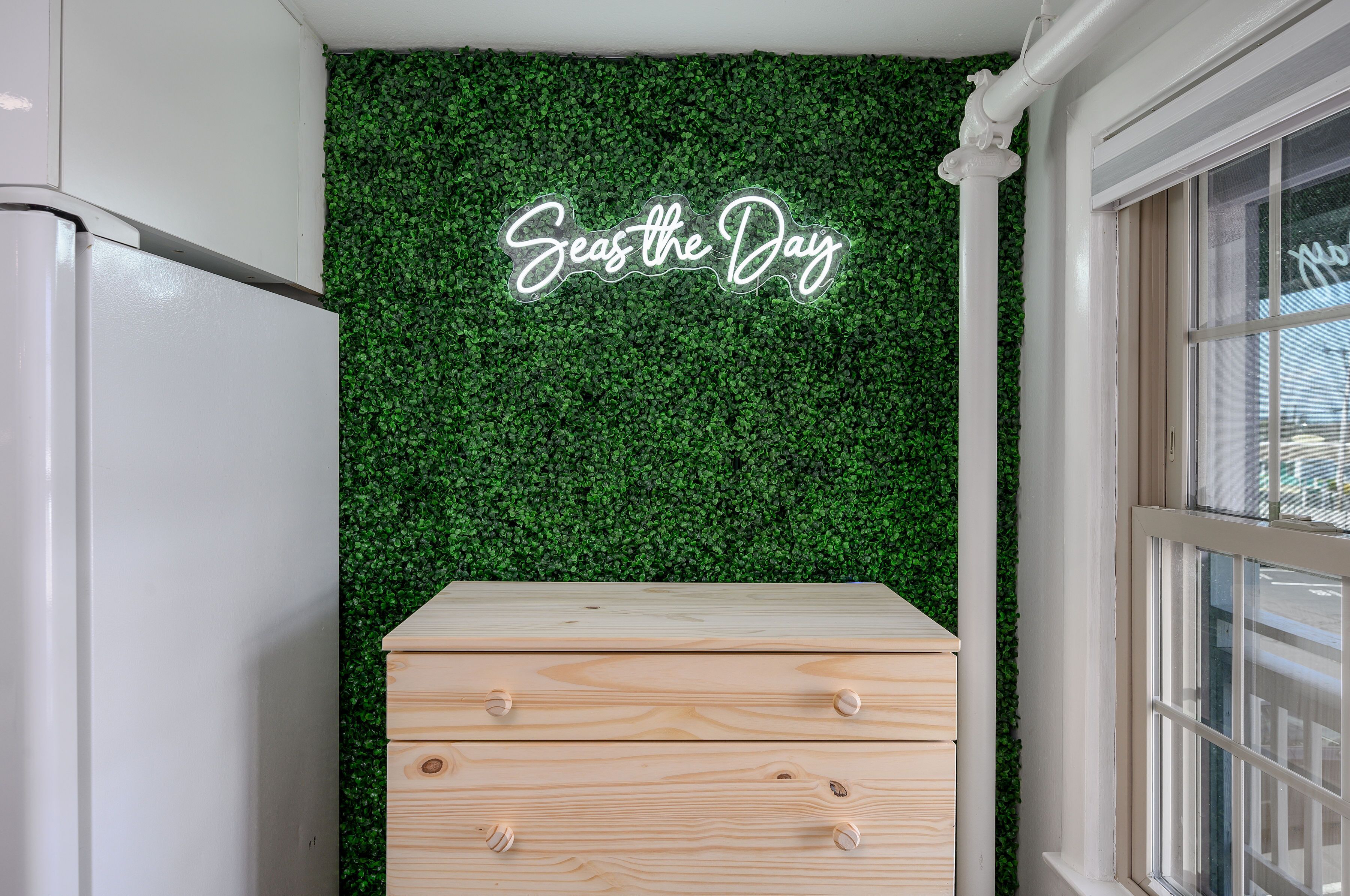 A room with a green artificial grass wall, a wooden chest of drawers, and a window with a view.