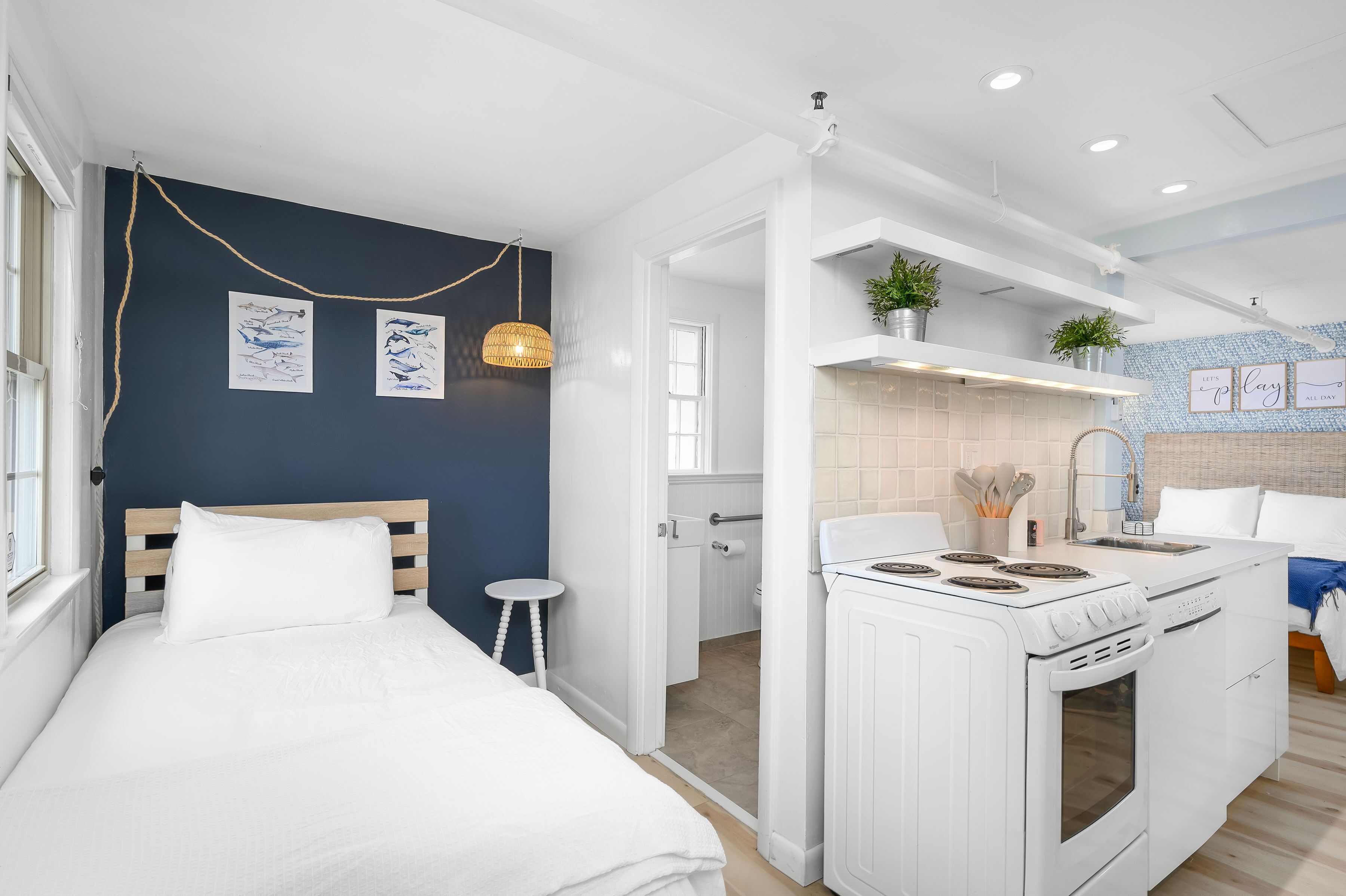 Ein modernes, kleines Zimmer mit einem Bett, einem kleinen Hocker, einer Küche inklusive Herd und einem durch eine offene Tür einsehbaren Badezimmer.