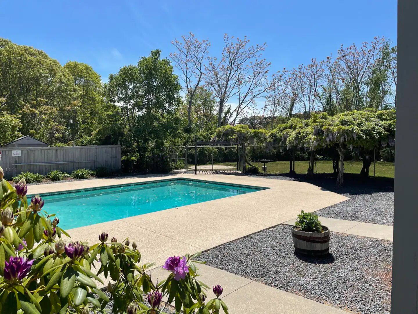 Una piscina rectangular con solárium de baldosas, rodeada por una zona de grava y una pérgola de madera.