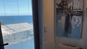 A room with a large sliding glass door leading to a balcony with a view of the ocean. There is a single chair with a fluffy pillow and a piece of abstract art on the wall. - Oakleigh House (Burnie)