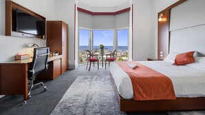 A hotel room with a large bed, a desk with a chair, a TV, and a view of the city through a large window. - Oakleigh House (Burnie)