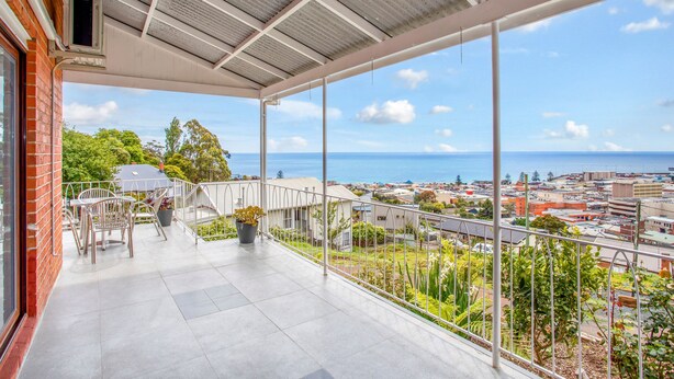 A balcony with a view of the sea and cityscape, featuring a table and chairs, and a potted plant. - Oakleigh House (Burnie)
