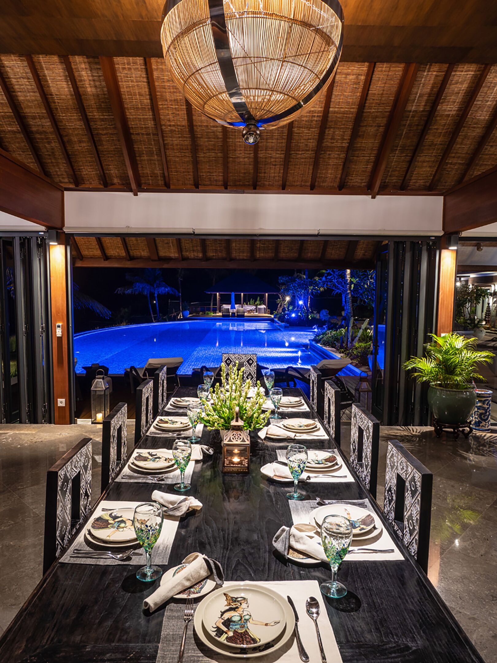 A dining area with a long table set for a meal, a decorative centerpiece, and a view of an outdoor swimming pool at night.