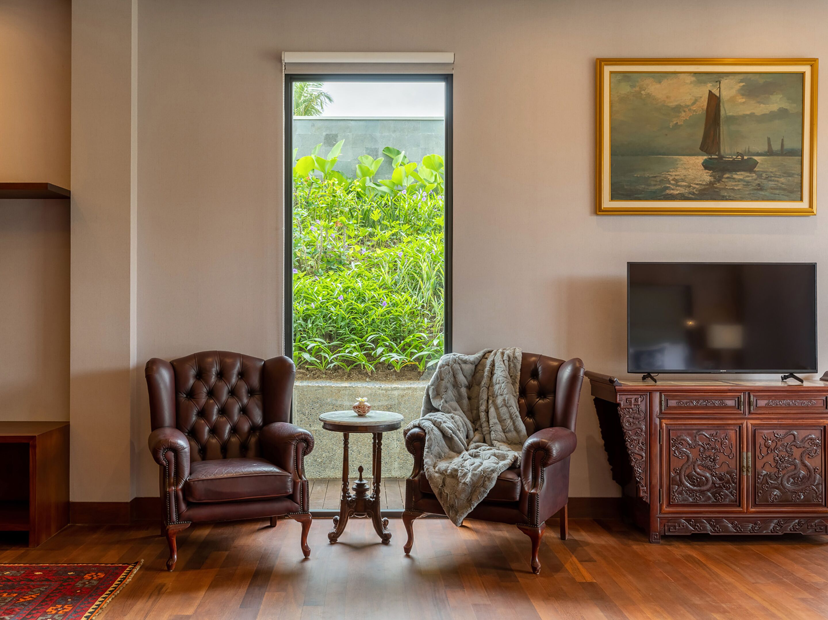 A room with two leather armchairs, a small round table, a television, and a cabinet with intricate carvings. A large window provides a view of greenery outside.
