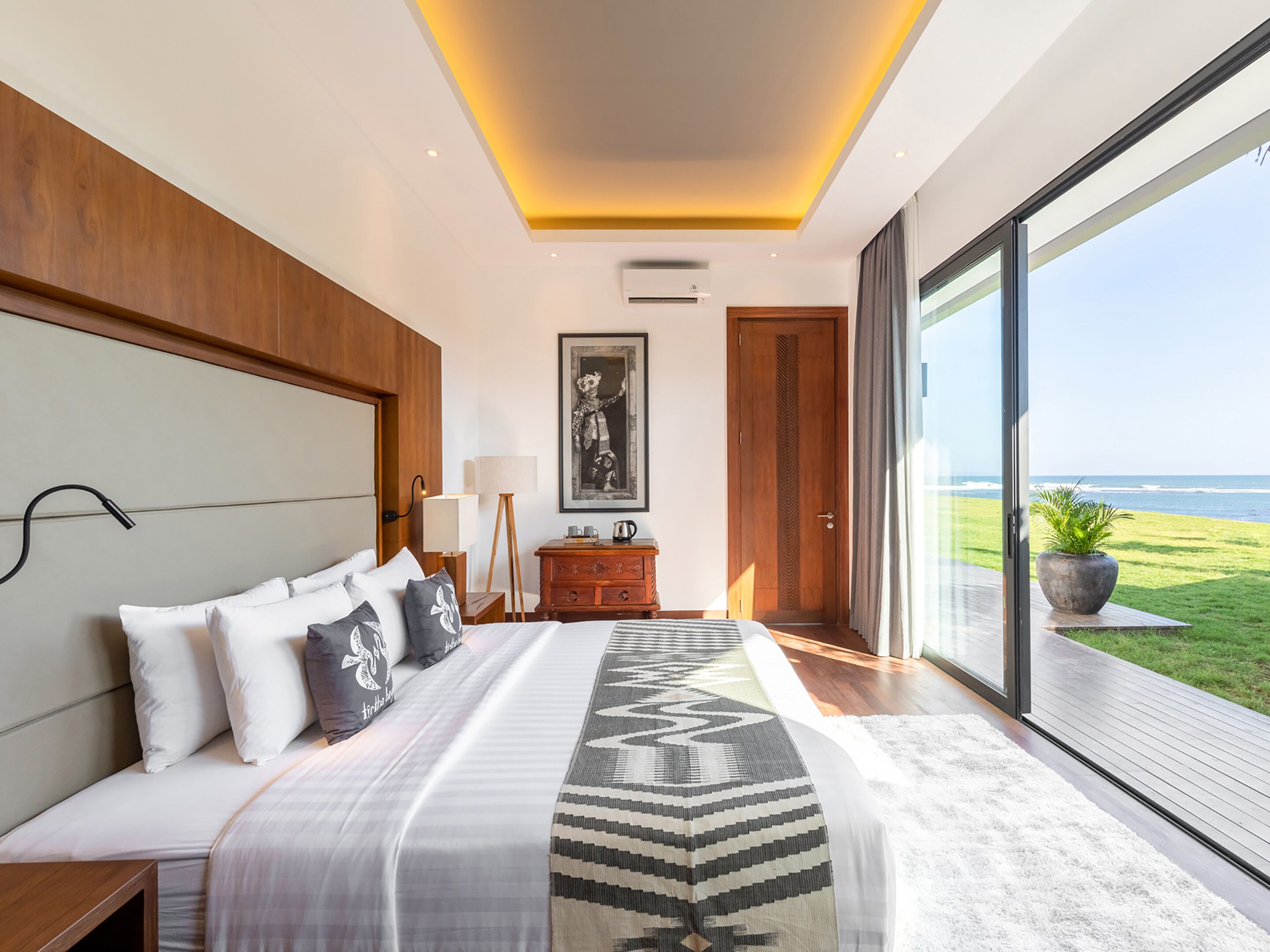 A modern bedroom with a large bed, a wooden headboard, a bedside table with a lamp, a dresser, and a large window offering a view of the ocean.