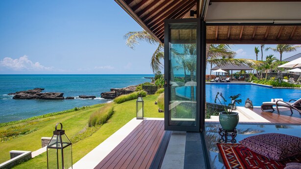 Una terraza moderna al aire libre con puerta de cristal que da a una zona de piscina, con vista a un océano azul y una costa rocosa.