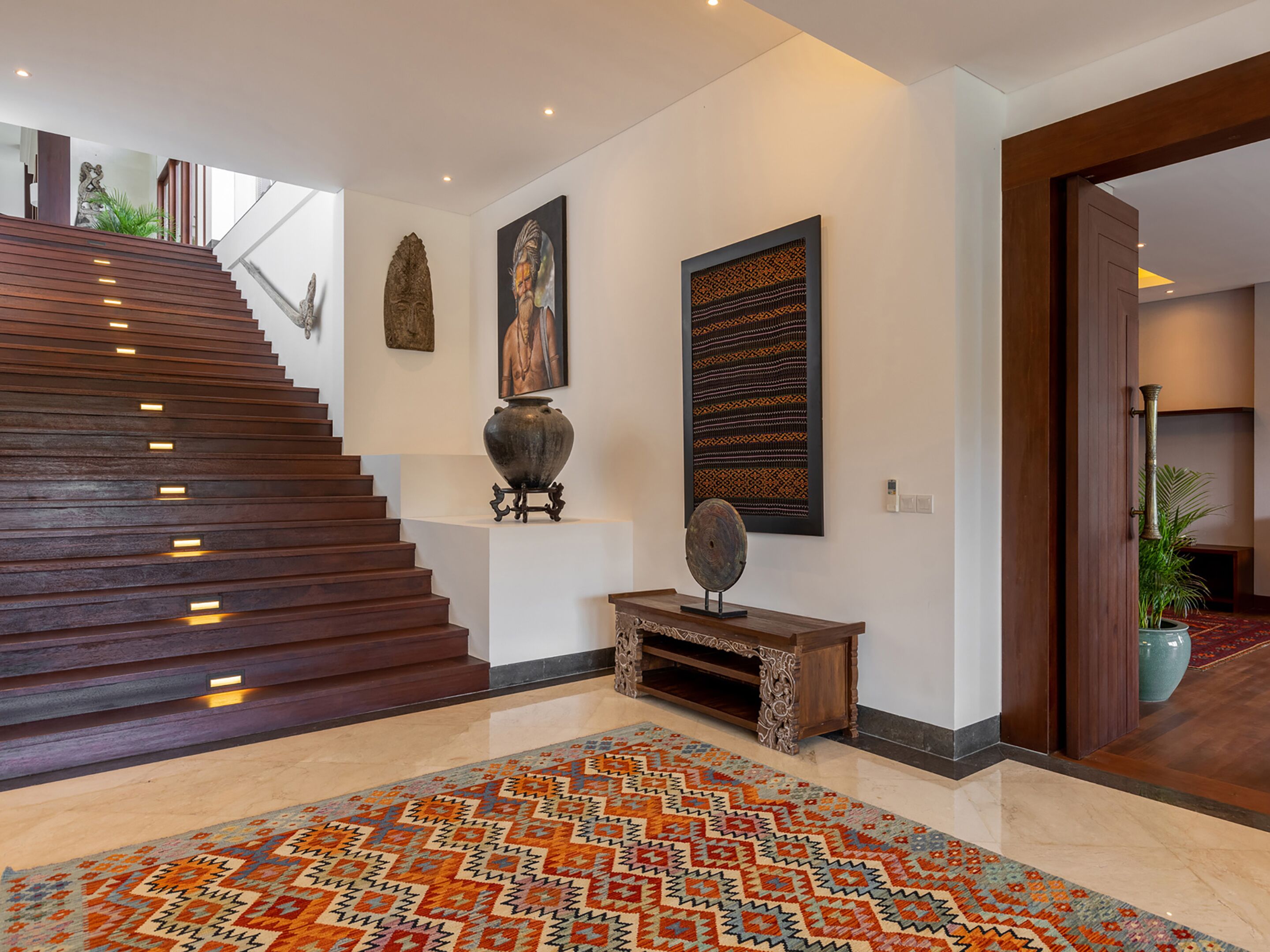 A modern interior with a wooden staircase, a patterned rug, and a large vase on a pedestal.