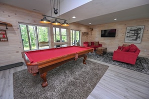 A pool table in a room with a red sofa, a television on the wall, and large windows.