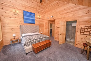 A wooden-paneled room with a bed, bedside table, bench, and a view of a forest through a door.