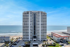 A high-rise hotel with a central entrance, located near a beach with a parking lot and a smaller building to the right.