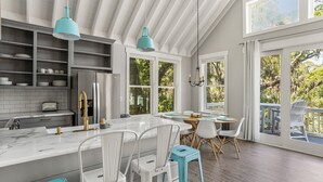 A modern kitchen with white cabinetry, a marble countertop, and a stainless steel refrigerator. There is a dining area with a wooden table and white chairs.