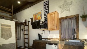 Una cocina compacta con armarios de madera, microondas y refrigerador. Se ve una escalera apoyada en la pared y una ventana pequeña con cortinas.