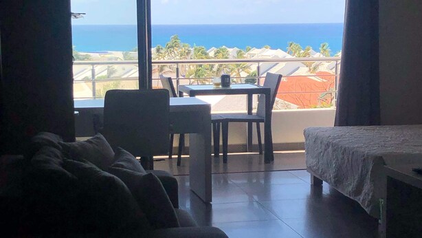 Habitació amb llit, sofà, taula i cadires. Hi ha un gran finestral amb vista al mar i palmeres.
