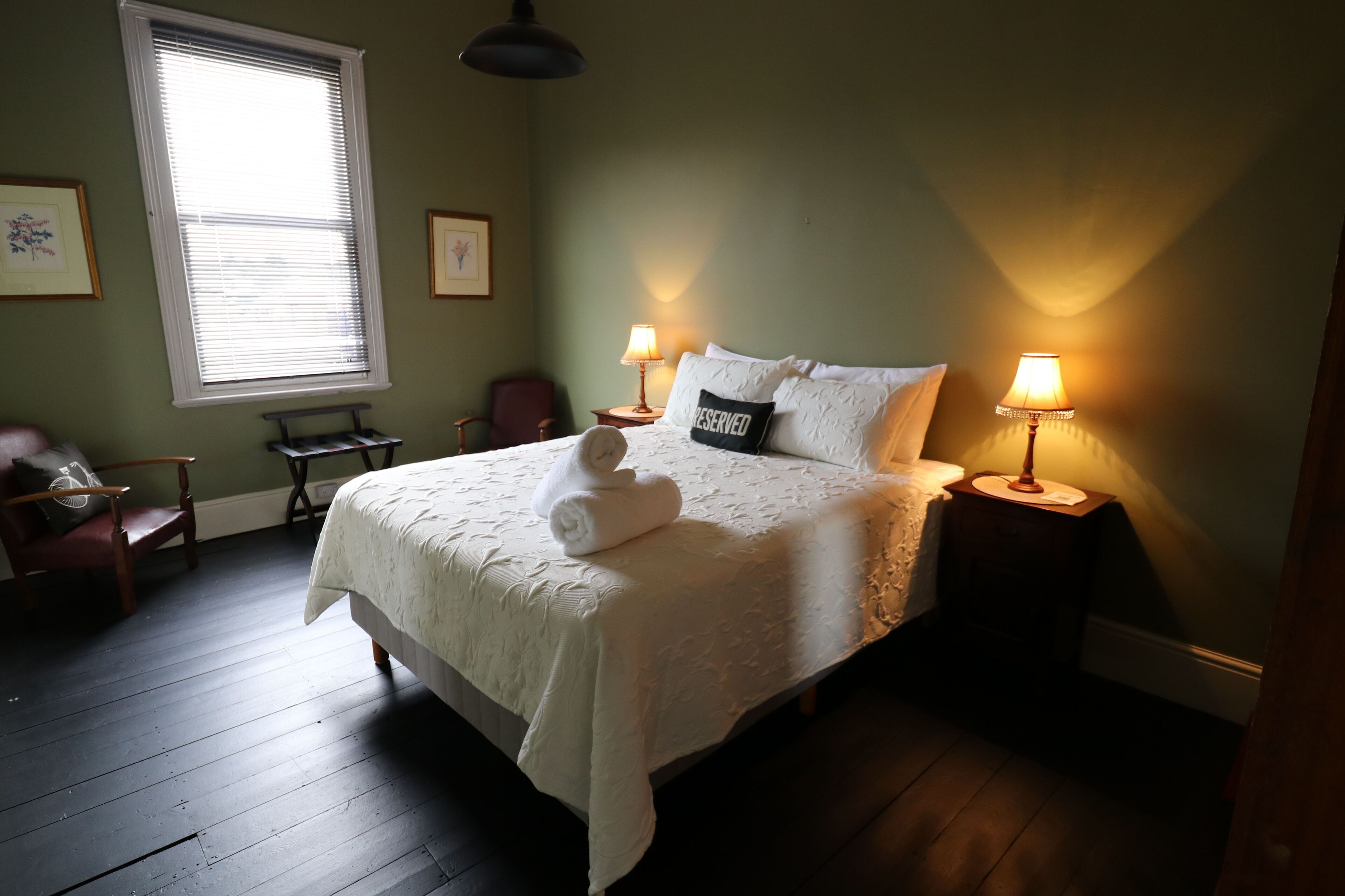 A neatly made bed with a white comforter and a towel, two bedside lamps, a chair, a small table, and a window with blinds.