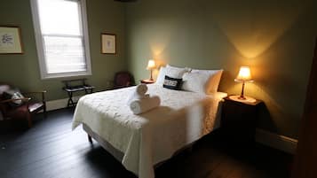 A neatly made bed with a white comforter and a towel, two bedside lamps, a chair, a small table, and a window with blinds.