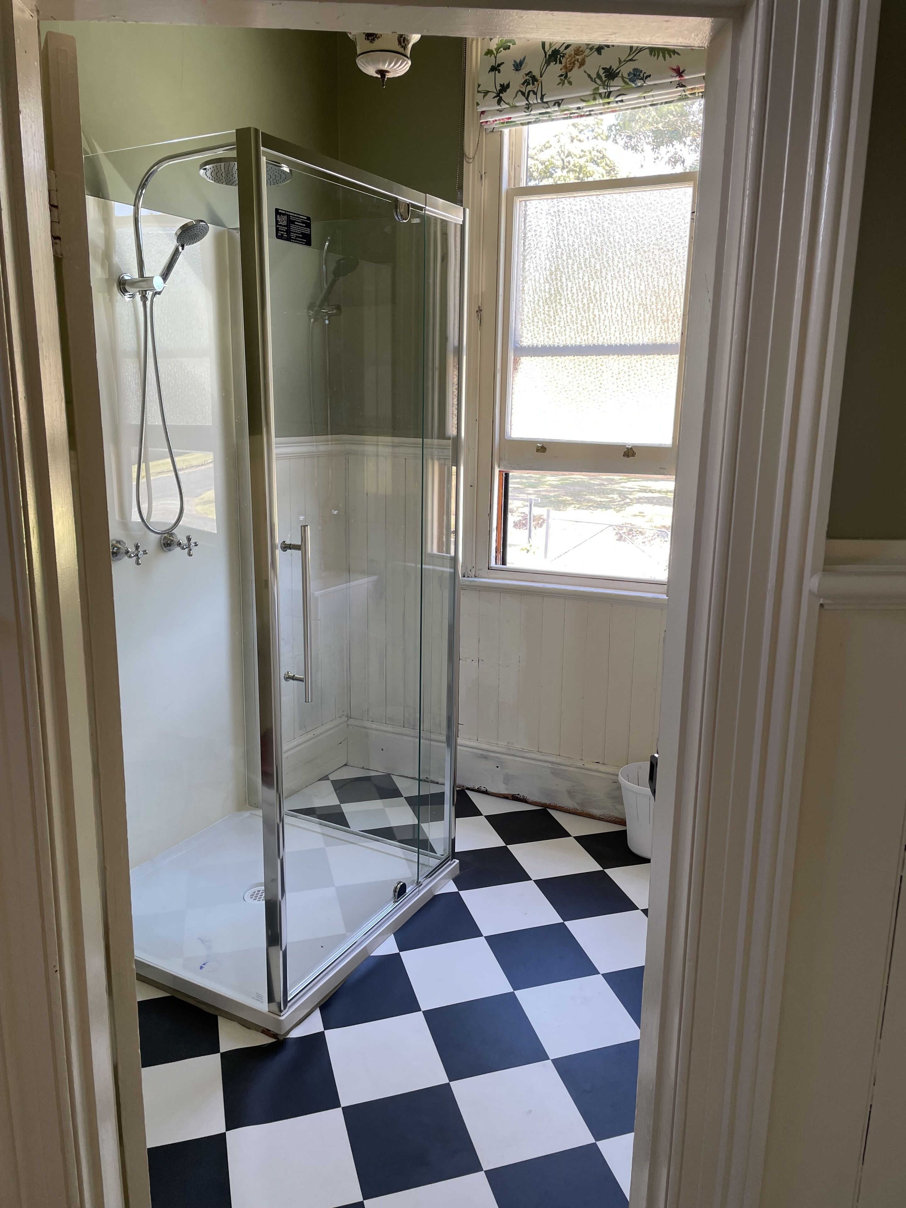 A glass-enclosed shower with a checkered floor, a window with a view, and a white door.