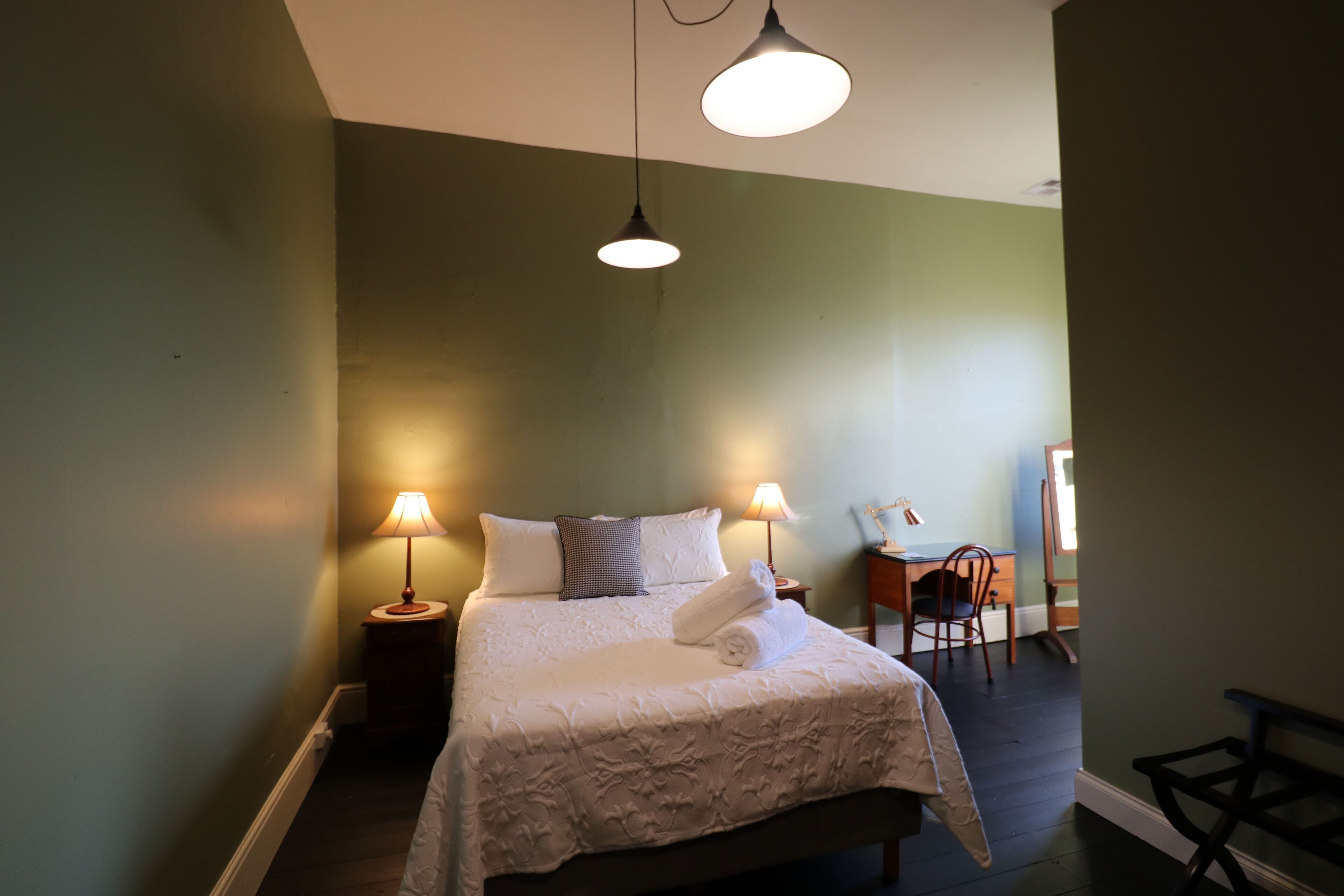 A neatly made bed with white linens and a rolled towel, two bedside lamps, a small wooden side table, and a chair in a room with green walls and dark wooden flooring.