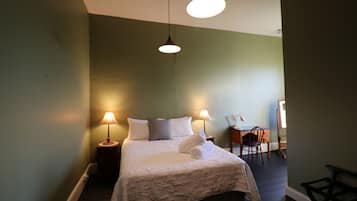 A neatly made bed with white linens and a rolled towel, two bedside lamps, a small wooden side table, and a chair in a room with green walls and dark wooden flooring.