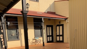 A wooden deck with a covered area, a building with a red roof, and a door with a black frame. - The Exchange Beaconsfield - Offering Heritage Accommodation (Beaconsfield)
