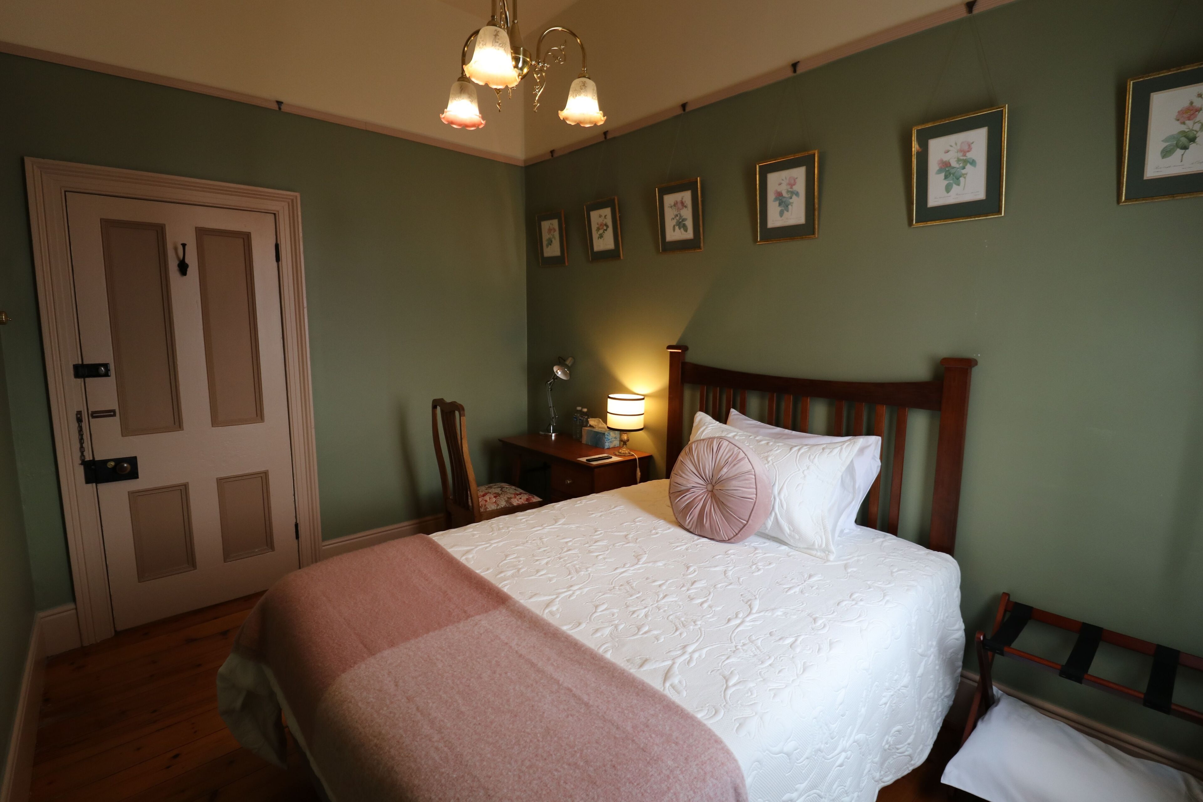 A neatly made bed with a white and pink bedspread, a wooden headboard, and a bedside table with a lamp. There are framed pictures on the wall and a door with a handle.