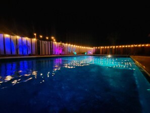 A nighttime swimming pool with illuminated vertical panels and string lights along the poolside.