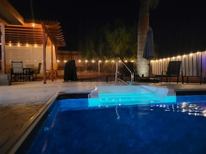 An outdoor swimming pool with a metal ladder, surrounded by chairs and a covered seating area with string lights.