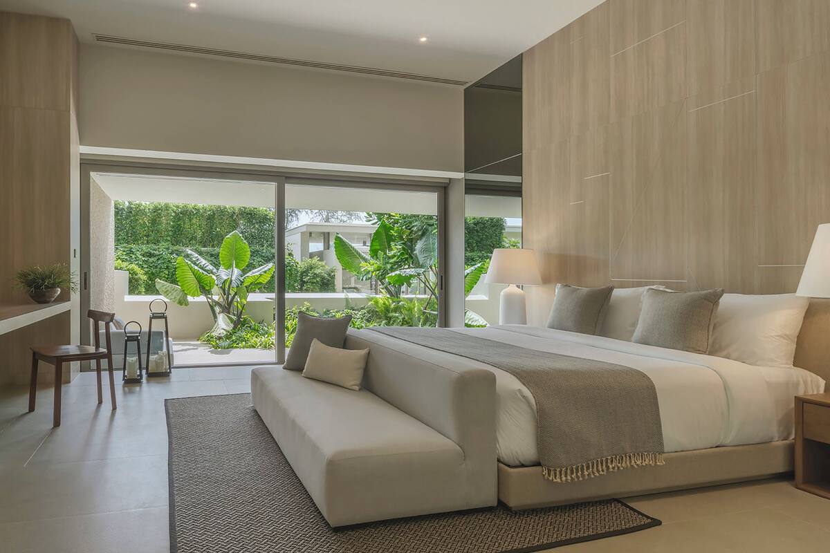A modern bedroom with a large bed, a sofa, a desk, and a view of greenery outside.