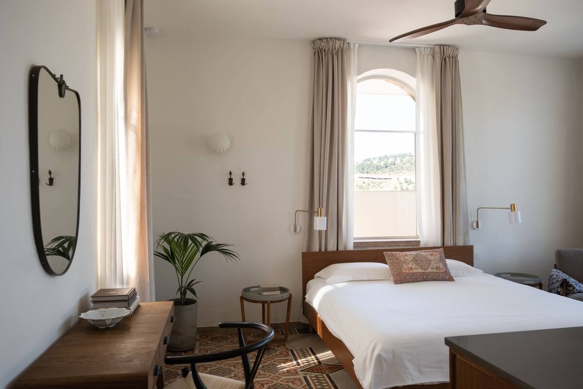 Une chambre d’hôtel bien aménagée, avec un lit, des tables de chevet, un bureau, une chaise, une plante en pot et une grande fenêtre avec des rideaux.