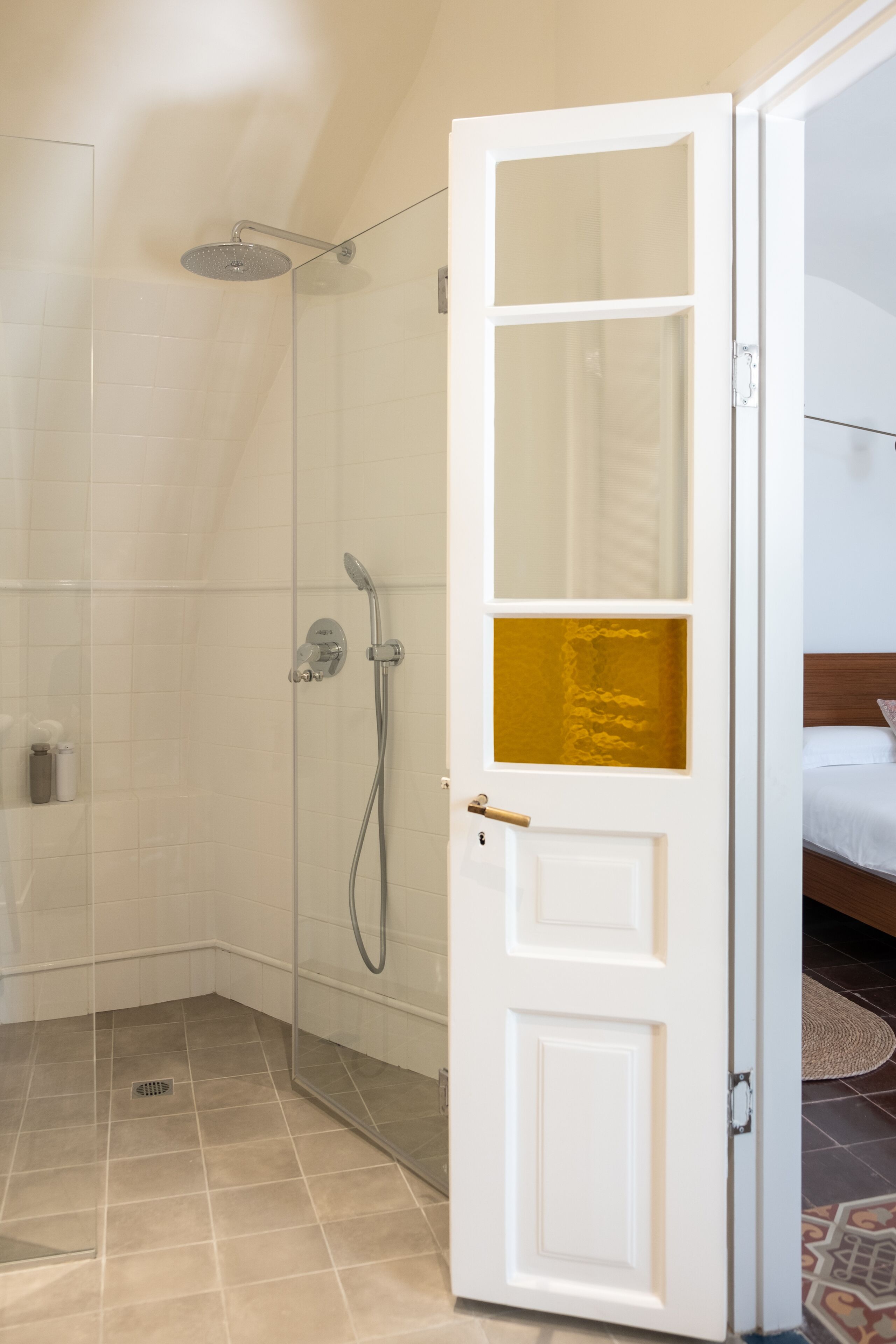 Un baño de hotel con ducha de vidrio, una puerta blanca con panel dorado y suelo de baldosas.