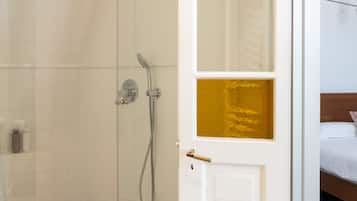 Un baño de hotel con ducha de vidrio, una puerta blanca con panel dorado y suelo de baldosas.