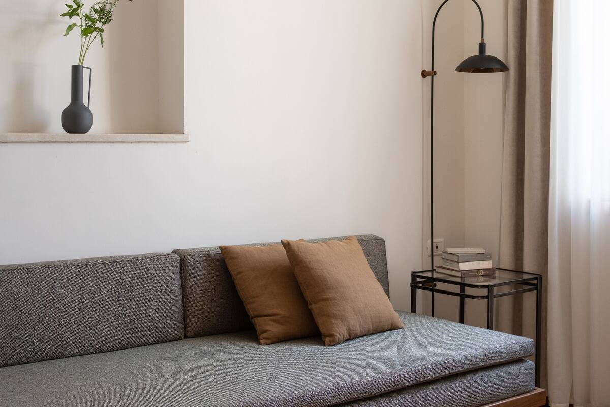 Un salon moderne avec un canapé gris, une petite table d’appoint avec des livres, une lampe sur pied et une plante en pot posée sur une étagère.