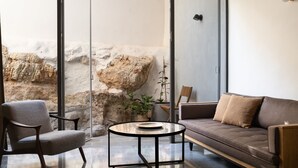 A modern living room with a glass coffee table, a sofa, and a chair. There is a large window with a view of a rocky wall. - KHAN EIN KAREM BOUTIQUE HOTEL (Jerusalem)