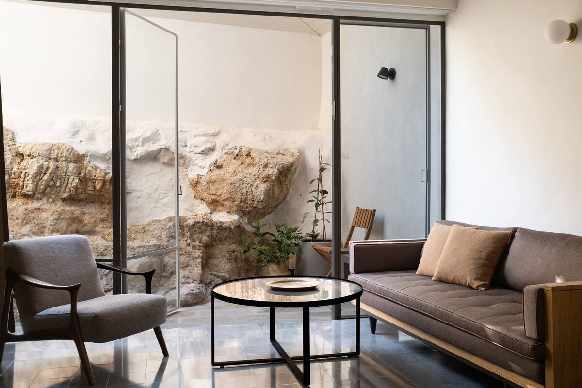 Un salon moderne avec une table basse en verre, un canapé et un fauteuil. Une grande fenêtre offre une vue sur un mur rocheux.
