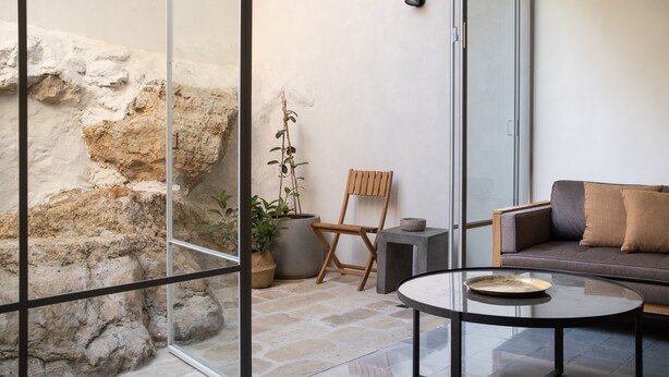 A modern living room with a glass partition, a stone wall, a wooden chair, a sofa, and a coffee table. - KHAN EIN KAREM BOUTIQUE HOTEL (Jerusalem)