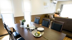 A dining area with a wooden table set for four, surrounded by chairs. There is a sofa in the background, and the room has a modern design with a neutral color palette. - Dynasty Nijuyonken (Sapporo)