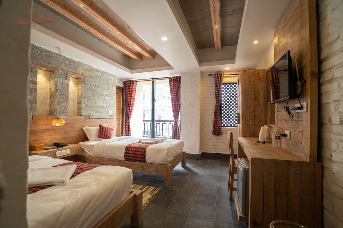 A hotel room with two beds, a wooden desk, a TV mounted on the wall, and a balcony with a view.