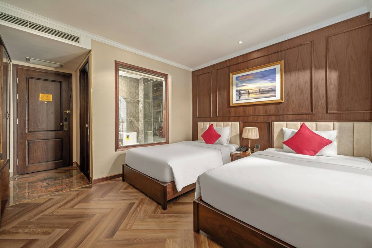 A hotel room with two beds, wooden flooring, a large window, and a painting on the wall.